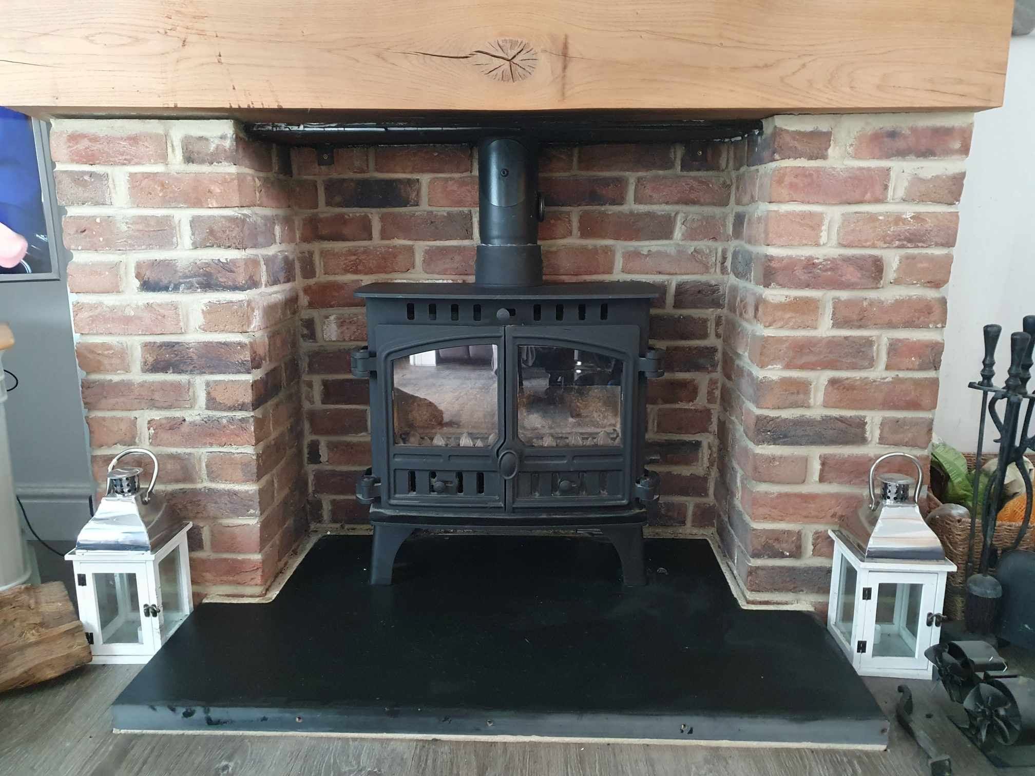 A black wood burner in an exposed-brick inglenook with a slate hearth and lanterns either side
