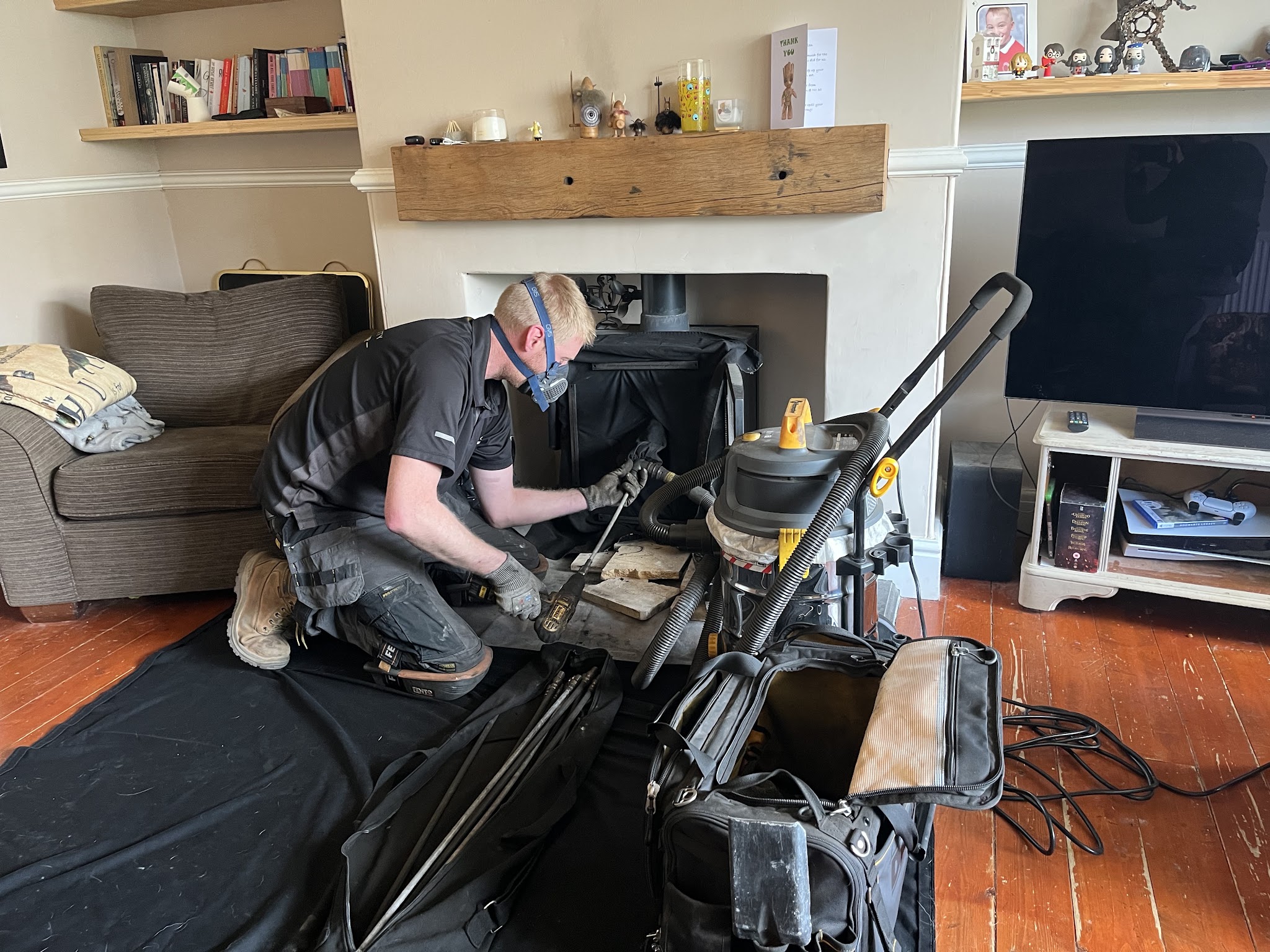 Arran at work on a wood burner in a living room, wearing a mask, with rods and vacuum on a black dust sheet
